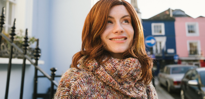 young person with red hair, wearing a knitted, colorful sweater, looking up and smiling outside in a city.